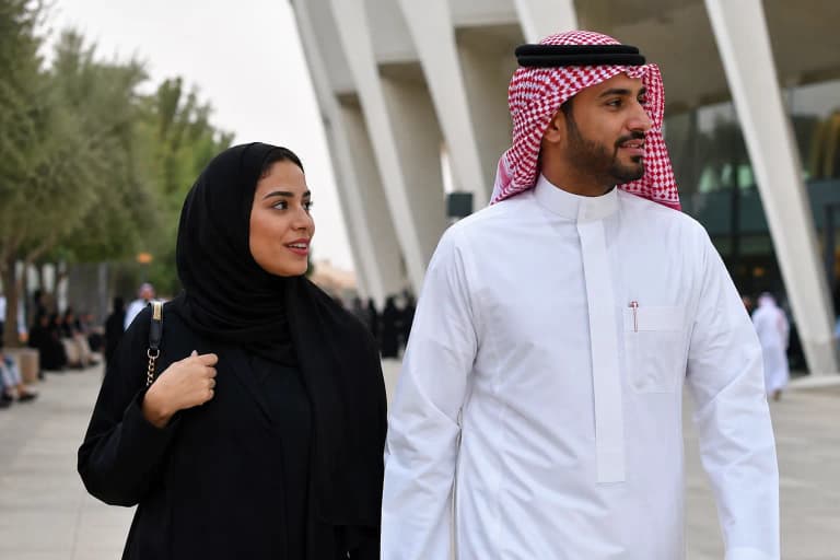 Man in white thobe with red ghutra headscarf and black agal, woman in flowing black abaya